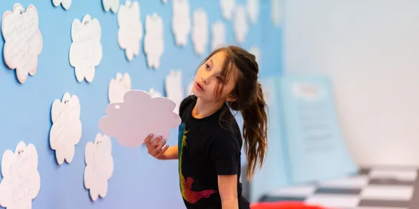 A child at an interactive learning exhibit, holding a cloud-shaped cutout next to a wall filled with similar cutouts.