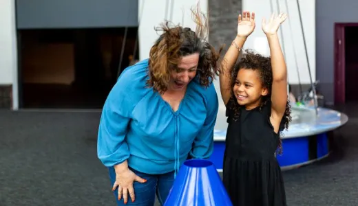 An adult and a child are stood in a science centre. The adult is putting their face over an air cannon and their hair is flying up. The Child is stood next to them with their hands in the air.