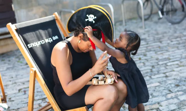 A small child reaching up to place a pirate hat on the head of an adult sitting near them who is trying to focus on a craft project at the same time.