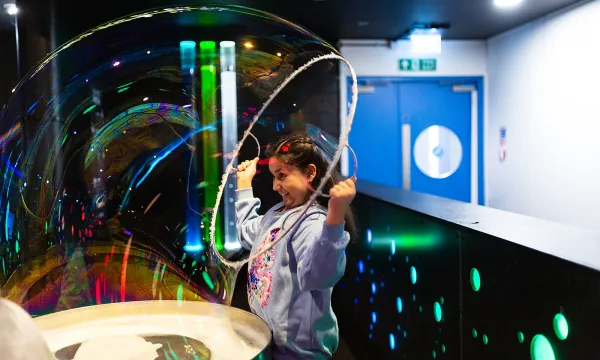 A child makes a large bubble using a large hoop which they are holding above their head.