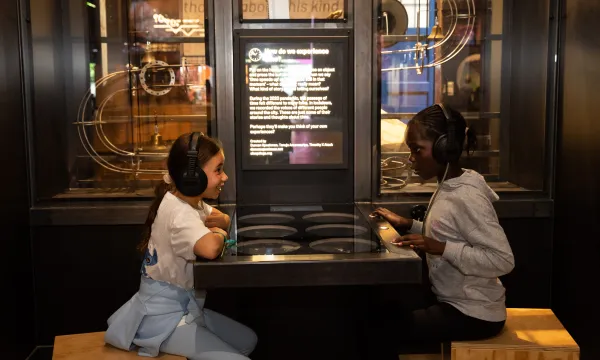 Two children are sat in a science museum opposite each other with a table in the middle of them. They are both wearing headphones 
