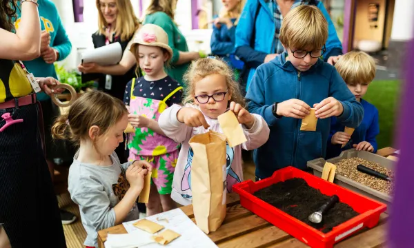An group of children are in a greenhouse playing with soil in plastic containers and paper bags.
