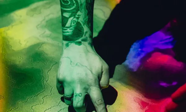 A person's tattooed arm reaching out into some sand, which is covered in contour lines and rainbow-coloured lights 