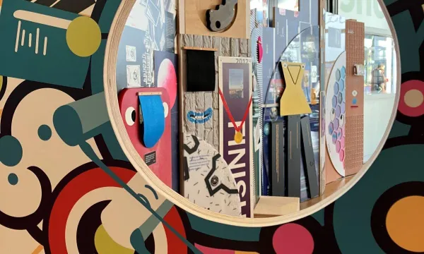 A round window in a colourful wall revealing an art display with geometric shapes, mirrors, and interactive elements in the foyer at We The Curious  