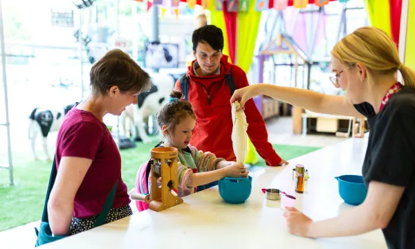 A child and two adults are stood behind a kitchen counter in a science museum. A member of staff is helping them squeeze oat milk from a cloth.