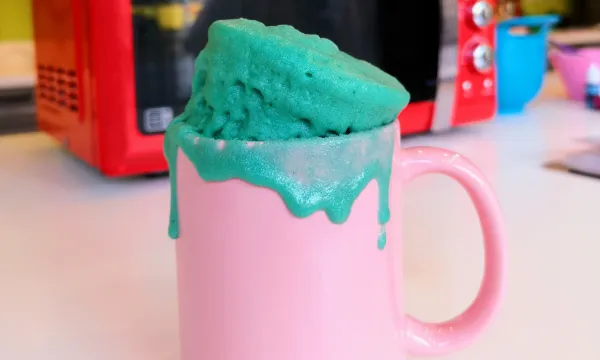 A pink mug is in front of a microwave with a bright blue mug cake spilling out of it onto the sides.