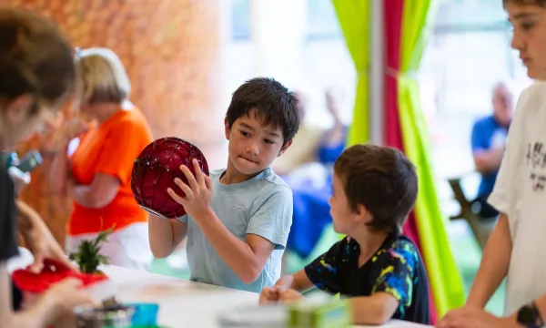 Two children are in the kitchen of a science centre. The older one is holding a big red ball and shaking it.