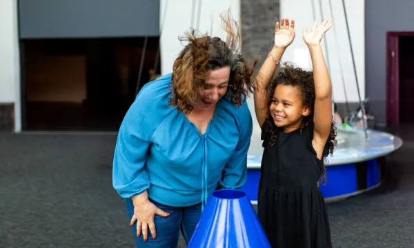 An adult and a child are stood in a science centre. The adult is putting their face over an air cannon and their hair is flying up. The child is stood next to them with their hands in the air. It's the Summer Holidays.