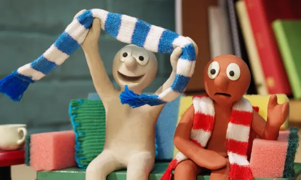 Two claymation characters are sat next to each other. One is holding a scarf and cheering and the other looks scared.