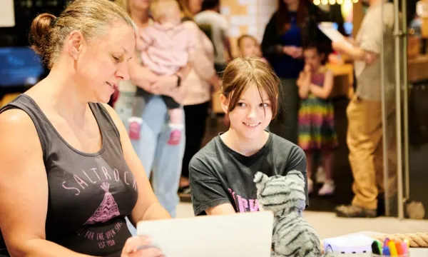 An adult and a child are sat down in a lab in a science centre. They are both wearing grey tops and on the desk is a white board, a snow leopard and a little robot.