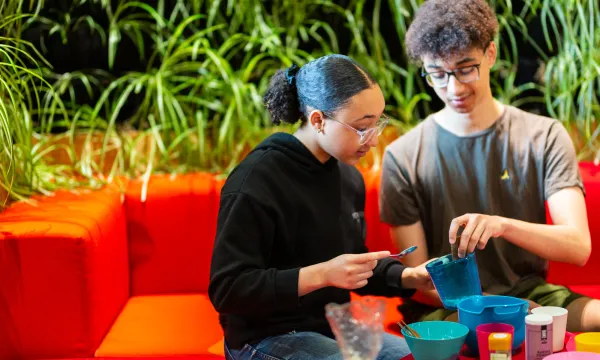 Two children are sat in the lab of a science centre on a red sofa. Looking down and playing with various kitchen equipment.