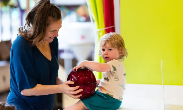 A child is seated on a counter top supported by an adult. They are holding a red sphere between them. 