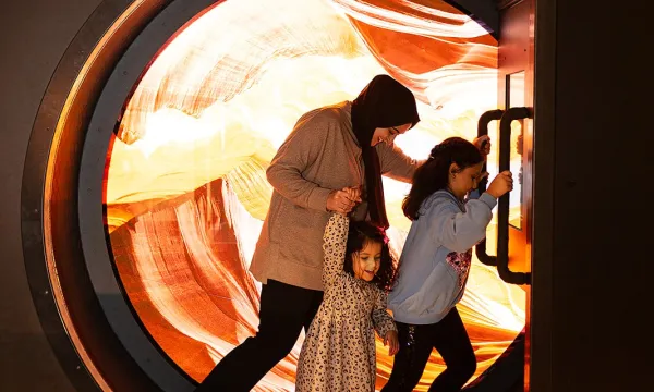 An adult and two children are shown in the wheel in the sand exhibit at We The Curious. There is a colourful red and yellow background inside the wheel.