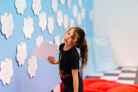 A child at an interactive learning exhibit, holding a cloud-shaped cutout next to a wall filled with similar cutouts.