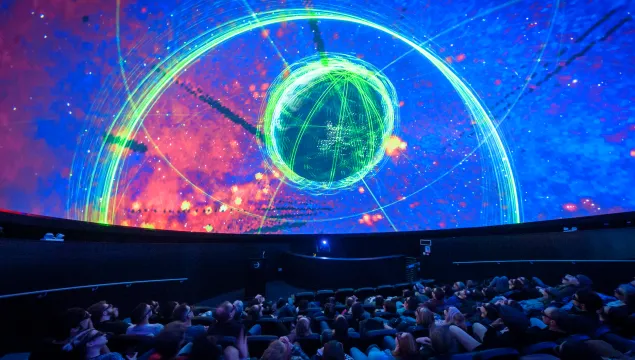 A shot of people sat in the Planetarium, a cinema-like experience. On the domed-shaped screen there's a visual of a colourful nebula.