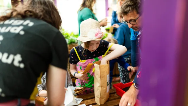 A child in a light sunhat and bright overalls putting seeds in a paper bag 