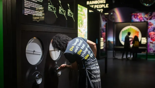 A member of We The Curious staff turns a handle on an exhibit. The question 'why do rainbows make people happy' is visible in the background. 