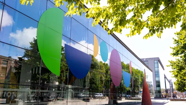 The glass exterior of We The Curious, which has large coloured letters spelling 'Curious'. Branches of a tree are visible in the foreground.