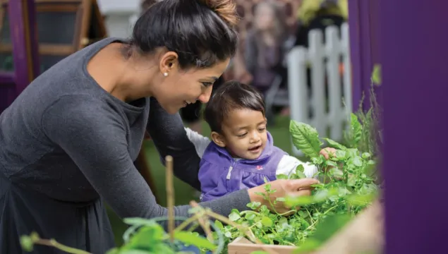 A person smiling and investigating plants with a toddler in the greenhouse exhibit
