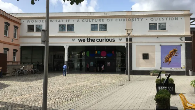 Large white building with 'We The Curious' written above the door, across a cobbled stone square