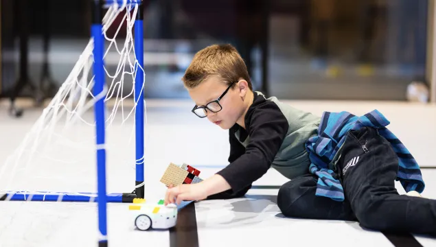 A child laying on the floor, pressing a small round wheeled robot, within a football goal