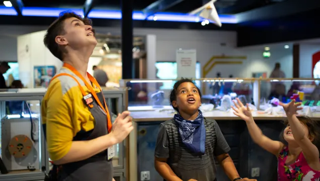 An adult and a child are in a science centre looking up at a piece of paper hovering in the air.