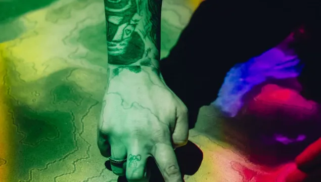 A person's tattooed arm reaching out into some sand, which is covered in contour lines and rainbow-coloured lights