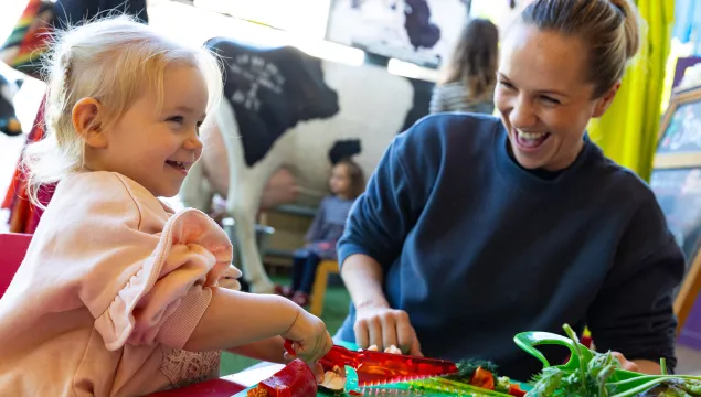 A young child and are smiling and interacting sat at a low table. The child is using a special plastic knife to cut a red pepper.