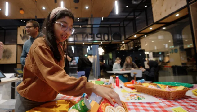 A child reaches for toy food set on a table with school dinner trays and bright gingham cloth.