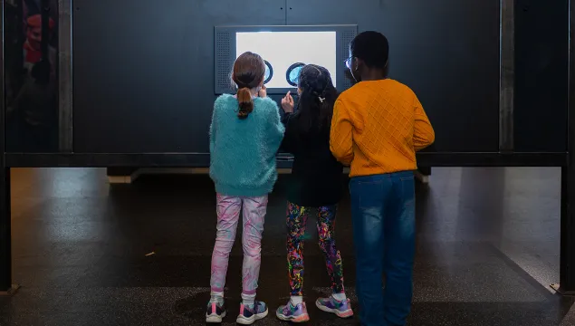 Three children look at an interactive exhibit through large magnifying glasses.