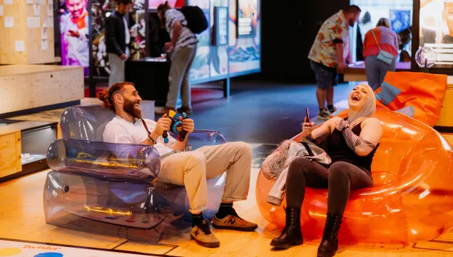 Two people are sat in the theatre of curiosity on bubble chairs, playing bopit and laughing.