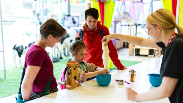 A child and two adults are stood behind a kitchen counter in a science museum. A member of staff is helping them squeeze oat milk from a cloth.