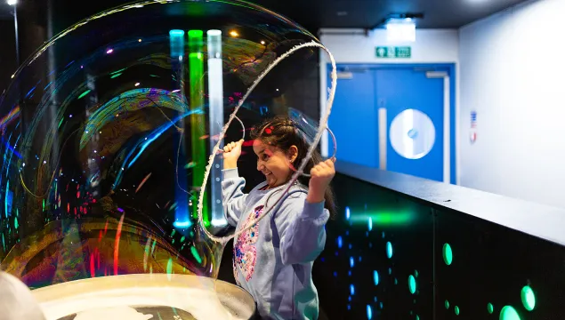 A child is in the middle of a science centre in a jumper pulling a sparkling bubble over their head.