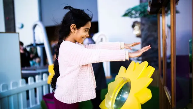 A child is stood in We The Curious science centre in a pink jumper. Stood Infront of a large, yellow, plastic flower and they are holding their hands up over it as it blows wind in their direction.