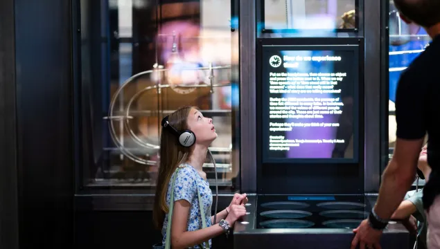 A child wearing headphones listens to stories in the Time exhibit at We The Curious. They are looking up at the exhibit.