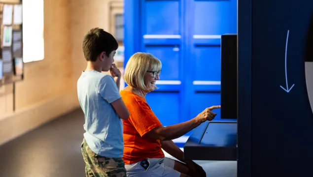 An adult and a child are in a science centre. The adult is sat down pointing at a screen and the child is stood next to them watching.