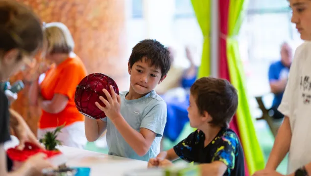 Two children are in the kitchen of a science centre. The older one is holding a big red ball and shaking it.