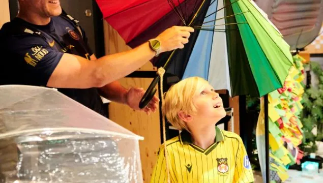A grownup opening a colourful umbrella over the head of a delighted looking child in an interactive station at We The Curious