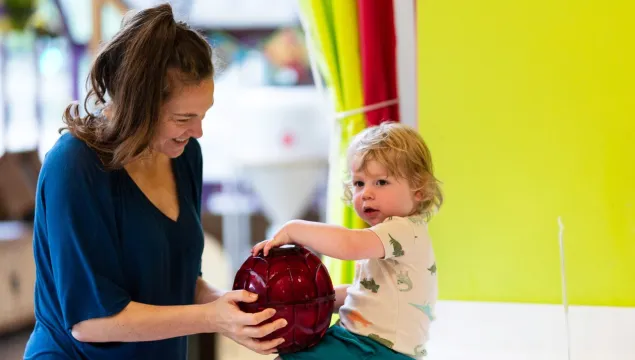 A child is seated on a counter top supported by an adult. They are holding a red sphere between them. 