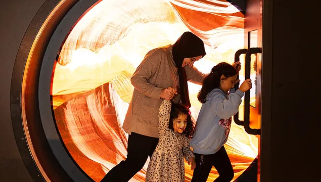 An adult and two children are pictured in the wheel in the sand exhibit at We The Curious. There is a colourful red and yellow background inside the wheel.