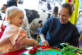 A young child and are smiling and interacting sat at a low table. The child is using a special plastic knife to cut a red pepper.