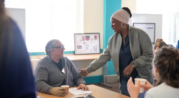 A person sat at a table with a coffee cup, with another person stood talking to them and touching their arm