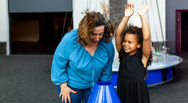 An adult and a child are stood in a science centre. The adult is putting their face over an air cannon and their hair is flying up. The child is stood next to them with their hands in the air. It's the Summer Holidays.