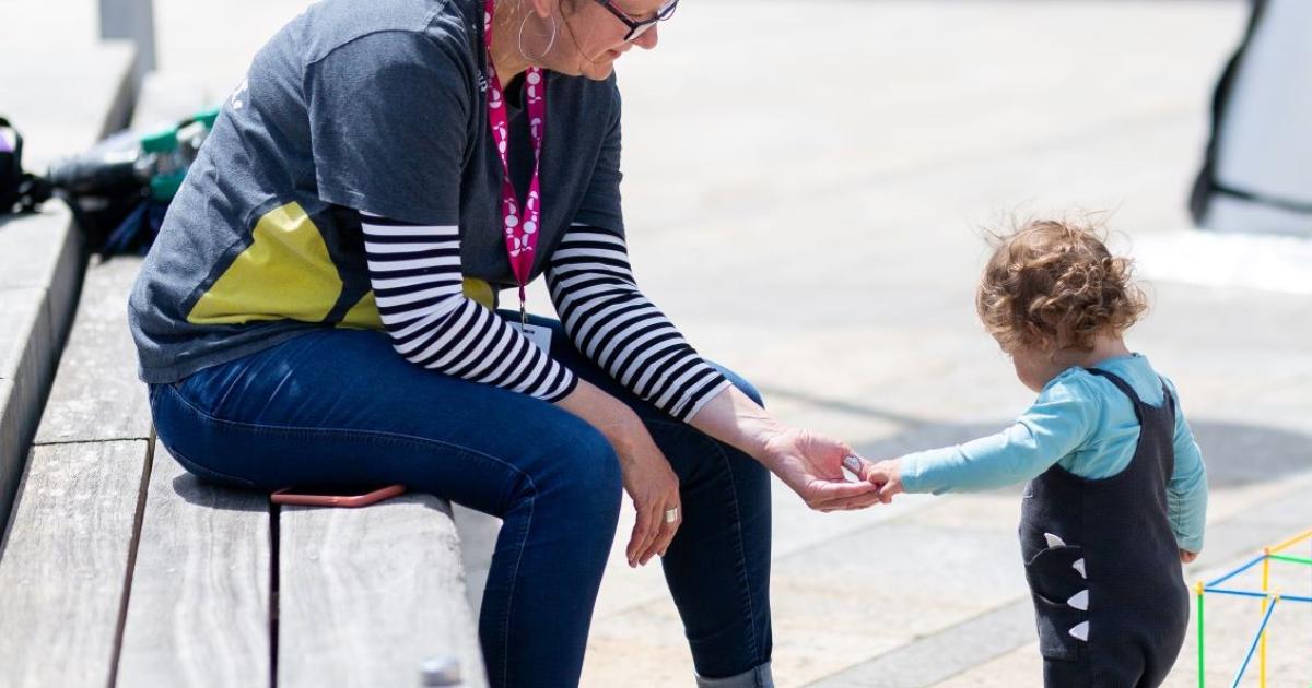 An adult and toddler playing with colourful straws together. The toddler is putting something in the adult's hand, they're outside in a stone paved space, with the adult sat on a wooden bench.
