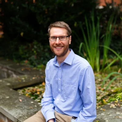 A person smiling with short brown hair and glasses, they are wearing a blue shirt and are sitting in front of some greenery.