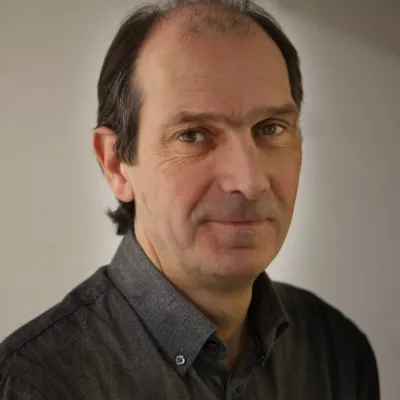 A person smiling with short brown hair, they are wearing a grey shirt, and are in front of a plain background.