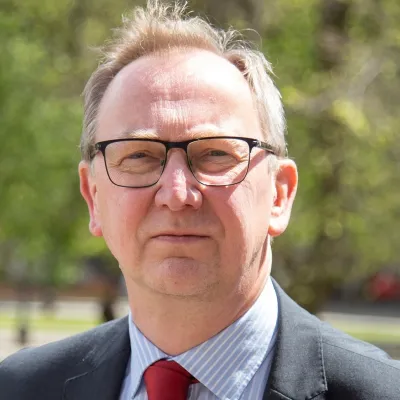 A person smiling with short brown hair and glasses, they are wearing a blue shirt, red tie and grey suit jacket, and are in front of some greenery.