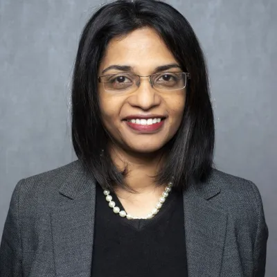 A person smiling with medium length black hair and glasses, they are wearing a black top and grey jacket, and are in front of a plain background.