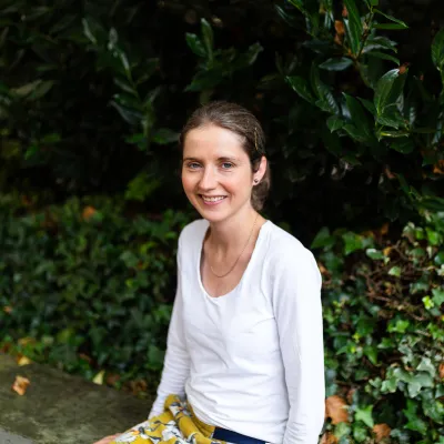 A person smiling with tied back brown hair, they are wearing a white top and are sitting in front of some greenery.