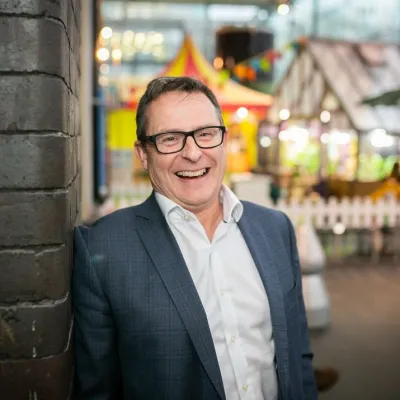 A person smiling with short brown hair and glasses, they are wearing a white shirt and grey suit jacket, and are standing in front of a We The Curious exhibit.